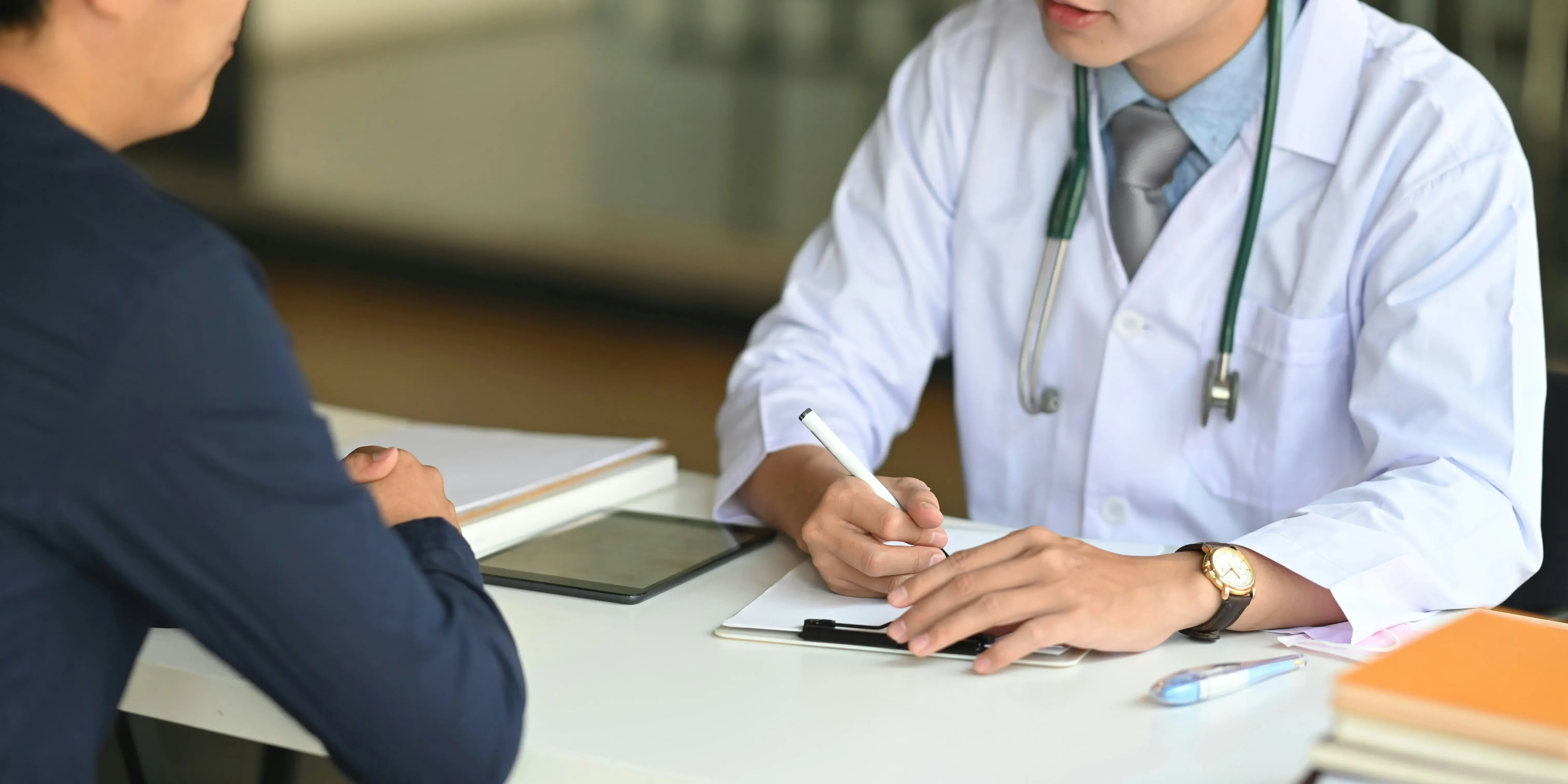 Läkare i vit rock som dokumenterar vid skrivbord tillsammans med patient.
